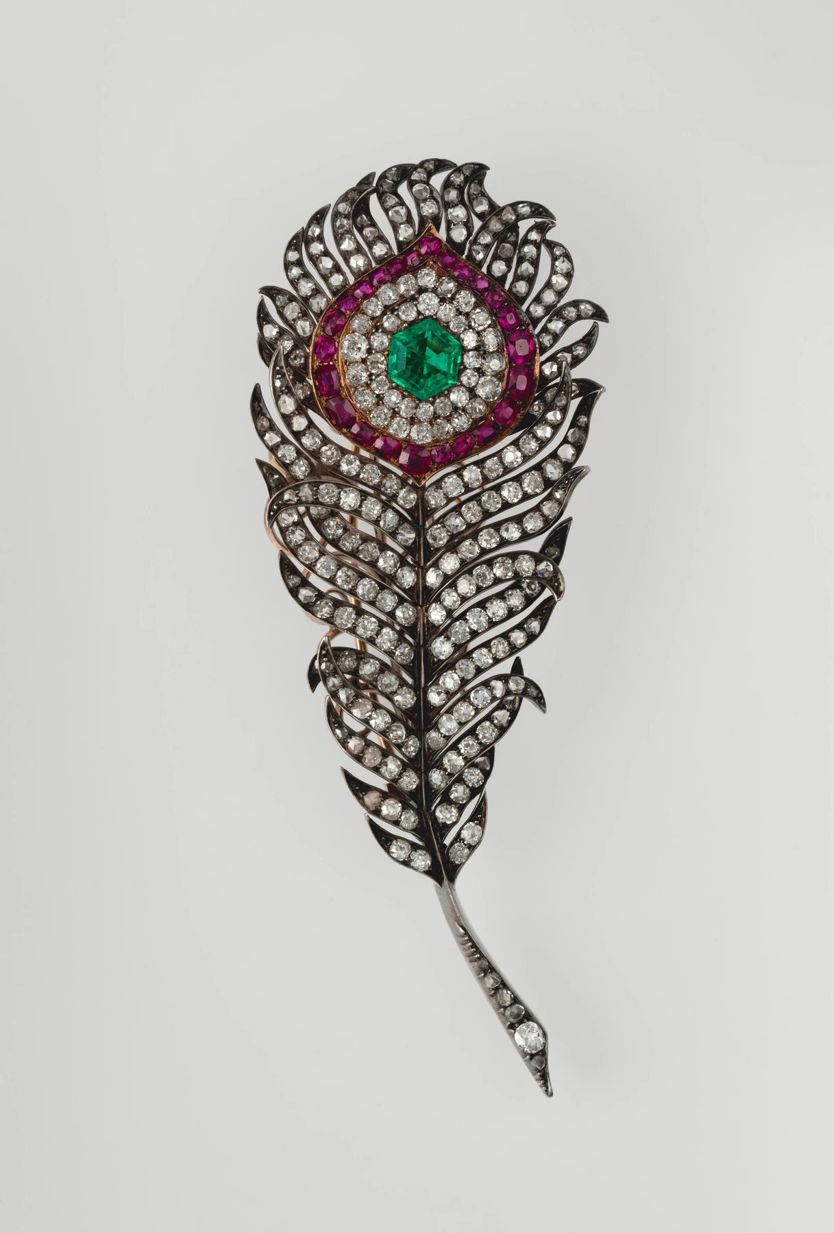 Broche histórico de pluma de pavo real con centro verde, orla roja y piedras claras.