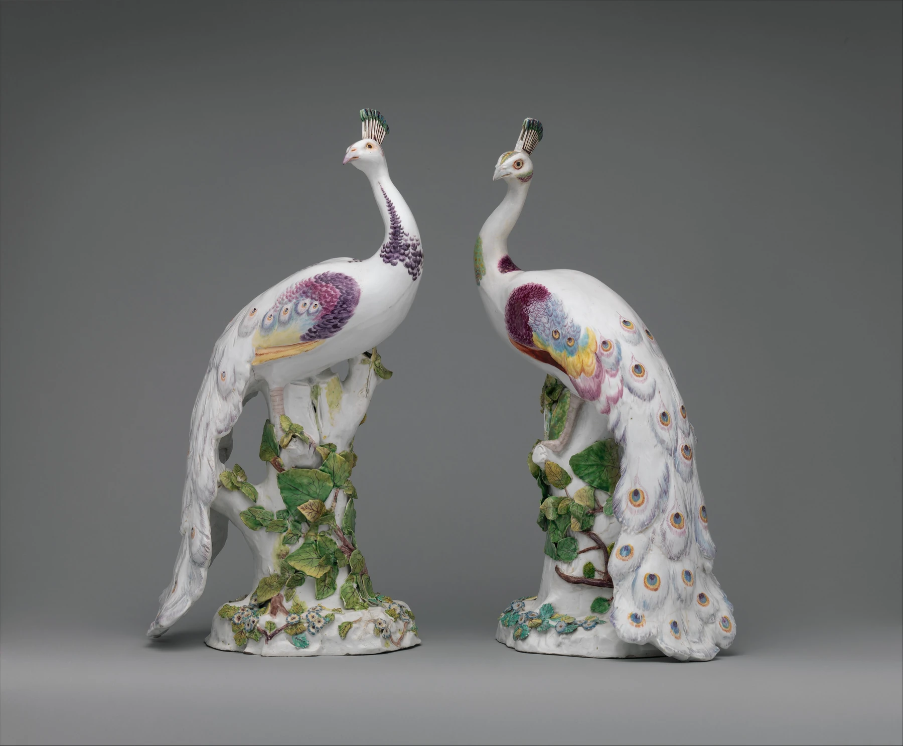 Dos figuras históricas de porcelana de pavo real sobre bases vegetales, con colas blancas decoradas.