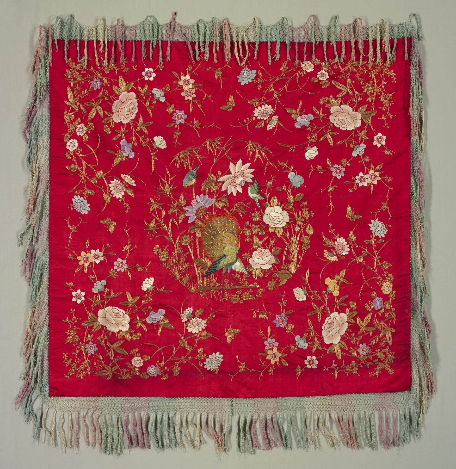 Mantel histórico rojo con flecos, bordados florales y un pavo real central.