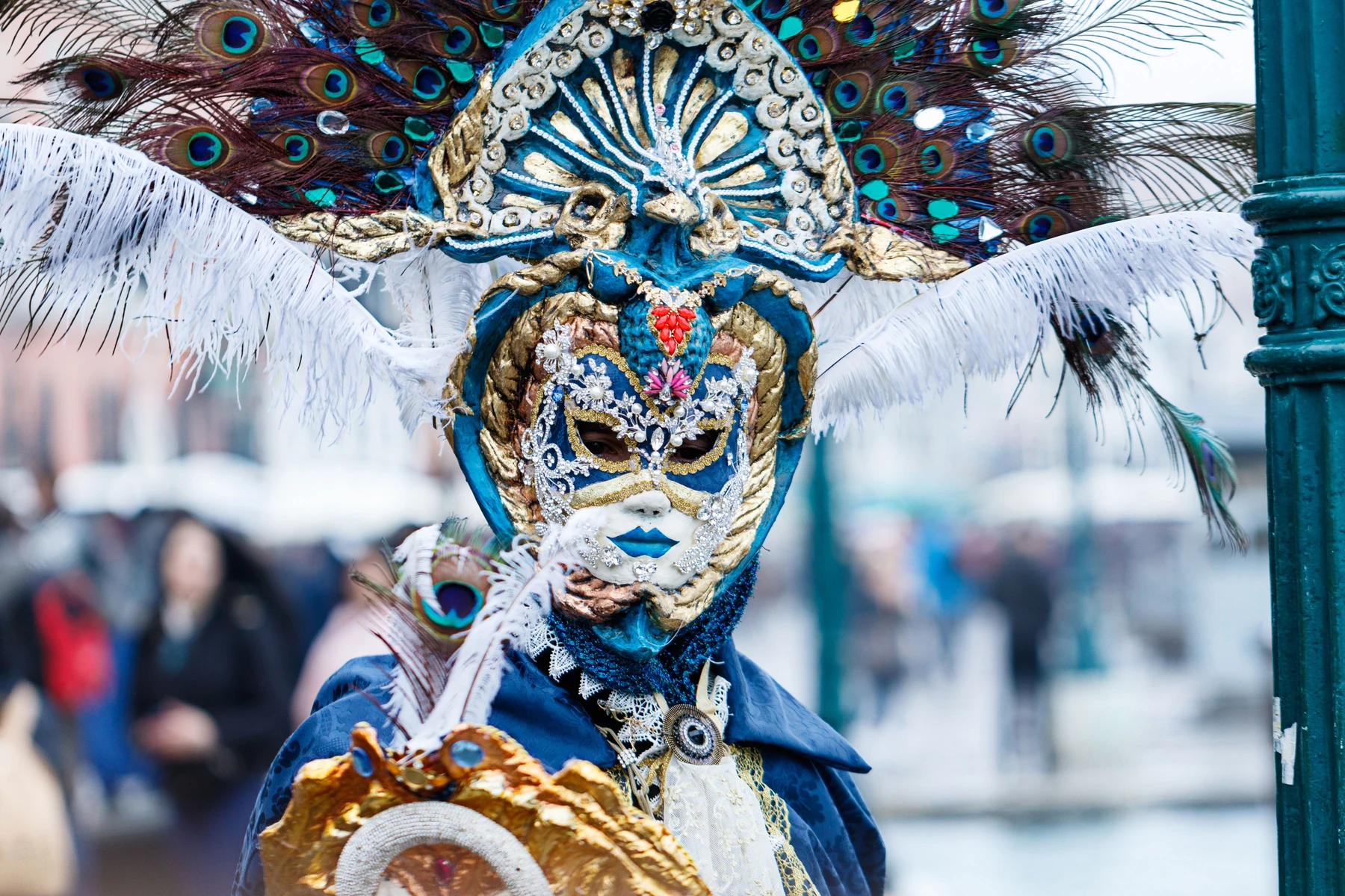 Máscara veneciana azul y blanca con tocado decorado con plumas de pavo real.
